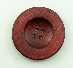 A close-up of a large, round, Red Wood Saucer Button from Buttons Etc. reveals its dark red hue with visible wood grain texture and four small holes in the center. Placed against a light background, this impressive button, with its 1 1/2" diameter, truly stands out.