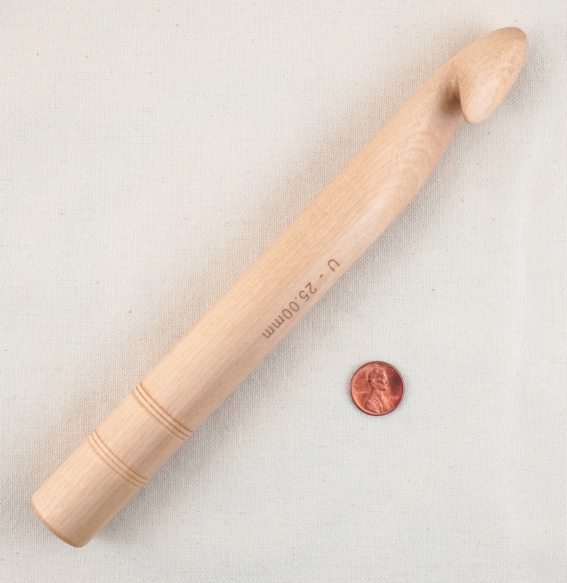 An Accessories Unlimited Basix Jumbo Crochet U (25mm) Hook crafted from wood rests beside a penny on a beige fabric background. Ideal for jumbo projects, this large crochet hook features a smooth, rounded handle adorned with two engraved rings near the bottom.