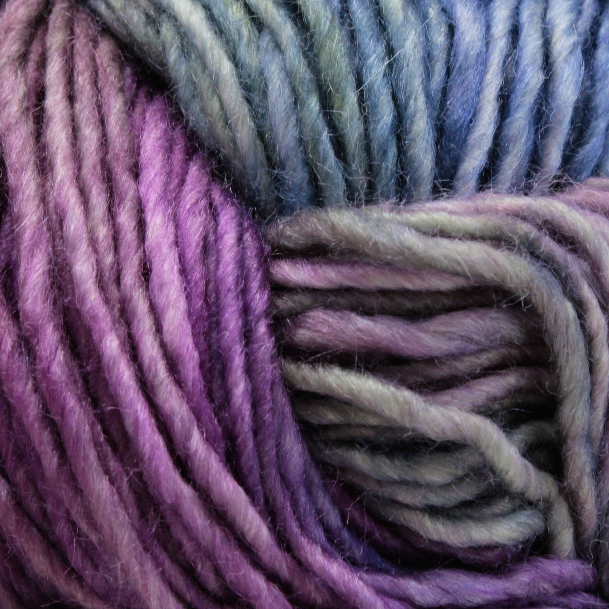 Close-up image of Malabrigo Silky Merino by Malabrigo Yarn, showcasing a luxurious gradient color scheme that transitions from deep purple to lavender and then to shades of blue and green. The yarn appears soft and slightly fuzzy, with a thick, twisted texture reminiscent of fine merino yarn.