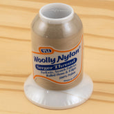 A spool of YLI Corp.'s Woolly Nylon Reinforcement Yarn stands upright on a wooden surface. The beige thread, ideal for knits, stretch fabrics, rolled hems, and edges, is labeled as made in Japan and 100% nylon. This versatile thread ensures security in your projects and even works well for crafting sturdy socks.