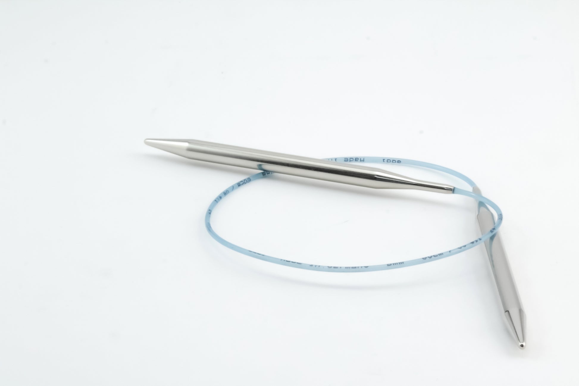 The Addi Turbo Circular Knitting Needles by Skacel are sleek, metallic tools with a distinctive design that mirrors short circular needles. They feature two tips connected by a flexible, light blue cord, which forms a loose loop as the knitting needles rest on a plain white background.