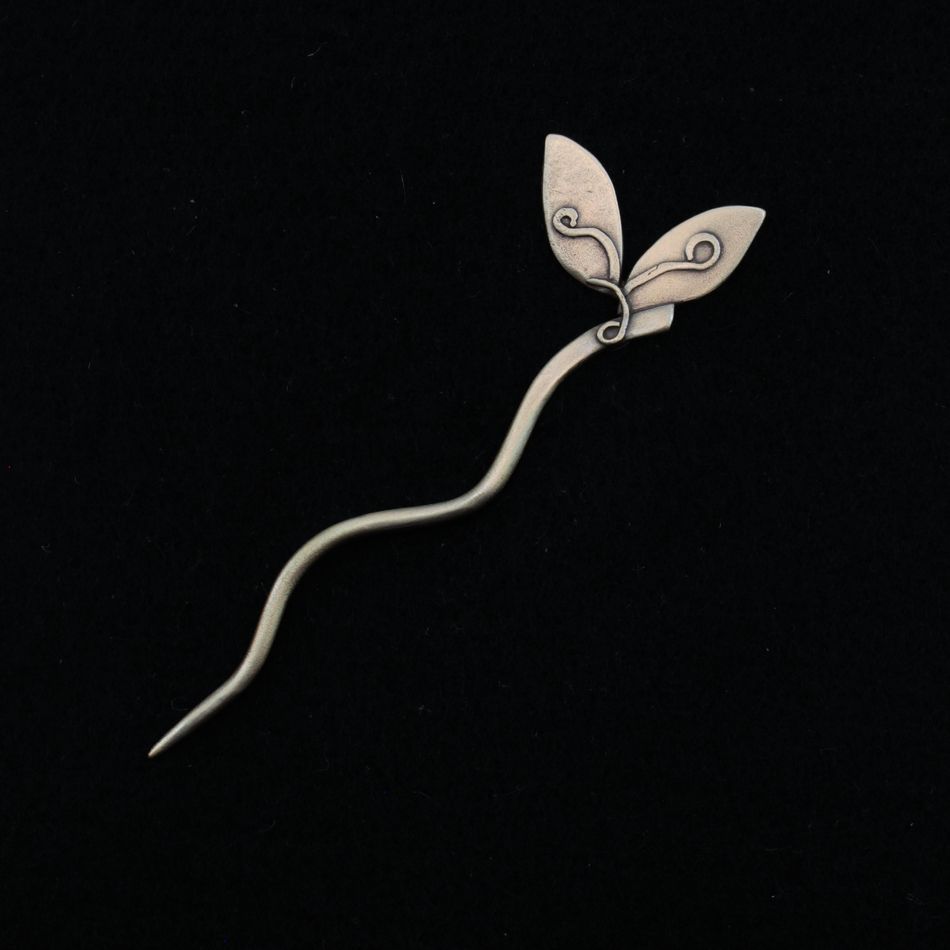 The Stick Shawl Pin by Bonnie Bishoff Designs, from the brand Bonnie Bishoff, features a silver hairpin with a long, wavy stem and two detailed leaves at one end. The leaves boast intricate swirl patterns. This handmade gift from a local Maine artisan is showcased against a solid black background.