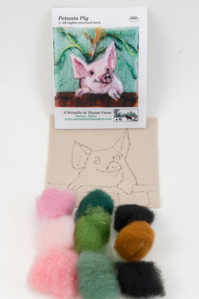 A Wrinkle in Thyme Farm's Thyme Tile Felting Kit (no tools) includes colored wool roving and a piece of fabric with a drawn outline of a pig. Behind the fabric is a reference card showing a picture of the completed project, "Petunia Pig," making it ideal for beginner felters.