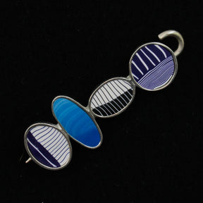 The Bubbles Shawl Pin by Bonnie Bishoff Designs is a decorative metallic object featuring four oval-shaped segments, each adorned with unique blue and white polymer clay-inspired patterns. The segments, connected in a straight line with a hook at the top, display various designs such as vertical lines, diagonal lines, and wave-like patterns set against a black background. This exquisite piece is crafted under the Bonnie Bishoff brand.