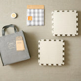 A flat lay image showcasing the Knitter's Block Kit by Cocoknits, featuring a grey tote bag with a tag, two interlocking beige foam mats from the kit, a grey and white checkered fabric pack for blocking handknits, and a small round container. All items are arranged on a beige surface.