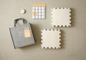 A flat lay image showcasing the Knitter's Block Kit by Cocoknits, featuring a grey tote bag with a tag, two interlocking beige foam mats from the kit, a grey and white checkered fabric pack for blocking handknits, and a small round container. All items are arranged on a beige surface.