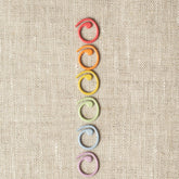 Six Cocoknits Split Ring Stitch Markers are arranged in a vertical line on a textured beige background. The objects are arranged in the following color order from top to bottom: red, orange, yellow, green, blue, and purple.