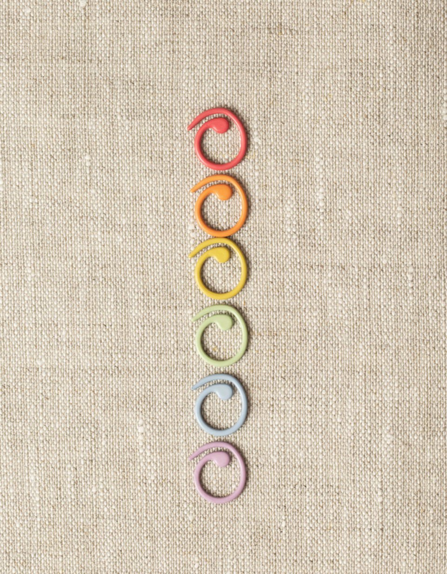 Six Cocoknits Split Ring Stitch Markers are arranged in a vertical line on a textured beige background. The objects are arranged in the following color order from top to bottom: red, orange, yellow, green, blue, and purple.