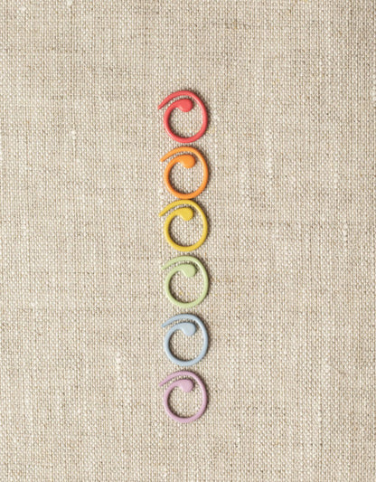 Six Cocoknits Split Ring Stitch Markers are arranged in a vertical line on a textured beige background. The objects are arranged in the following color order from top to bottom: red, orange, yellow, green, blue, and purple.