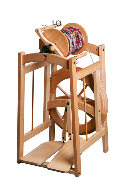 The Ashford Country Spinner 2 Spinning Wheel, created by Ashford Handicrafts Limited, is a wooden spinning wheel equipped with a foot pedal at the base for spinning yarn. It features multiple components, including a bobbin that holds multicolored textured yarn. The frame has a natural wood finish, providing it with a classic and rustic appearance.