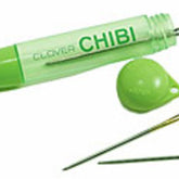 A green plastic case from Bryson Distributing, Inc. labeled "Chibi 3-needle Tapestry Needle Assortment" with its matching cap removed reveals a sewing needle inside. Two additional needles, one gold and one silver, lie beside the case, completing this handy set of three tapestry needles.