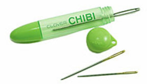 A green plastic case from Bryson Distributing, Inc. labeled "Chibi 3-needle Tapestry Needle Assortment" with its matching cap removed reveals a sewing needle inside. Two additional needles, one gold and one silver, lie beside the case, completing this handy set of three tapestry needles.