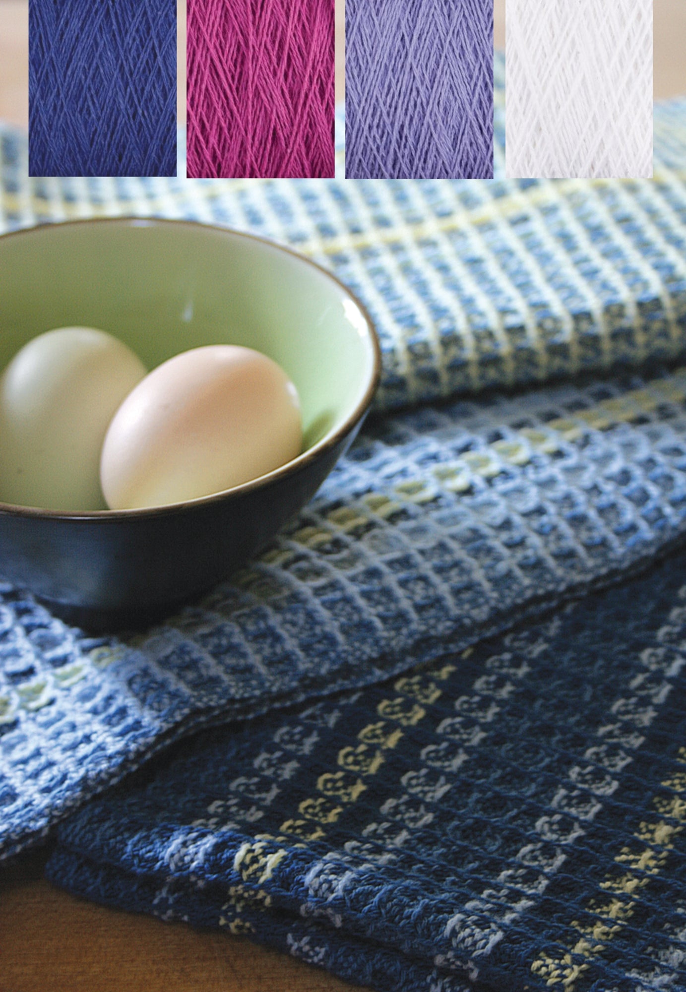 A green bowl containing two eggs is placed on the Waffle Weave Dish Towel Kit by Halcyon Yarn, which features various shades of blue. Above the bowl, there are four small swatches of yarn in blue, pink, purple, and white. The background is a wooden surface.