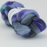 A large skein of Nube by Malabrigo is shown, featuring a mix of blue, purple, teal, and gray shades. The hand-dyed merino wool from Malabrigo Yarn is braided neatly and has a soft, fluffy texture. A label with text is partially visible wrapped around the wool. The background is plain white.