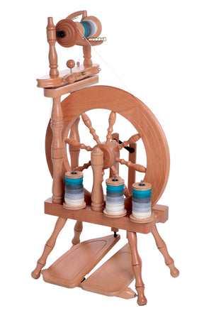 The Ashford Traveller Spinning Wheel, Double-Treadle by Ashford Handicrafts Limited is a traditional wooden spinning wheel with foot pedals, featuring spools of yarn in various shades of blue and white. The intricate design includes carved wooden details and a built-in lazy kate with multiple spindles for holding the yarn. This light, compact castle-style wheel is perfect for traveling.