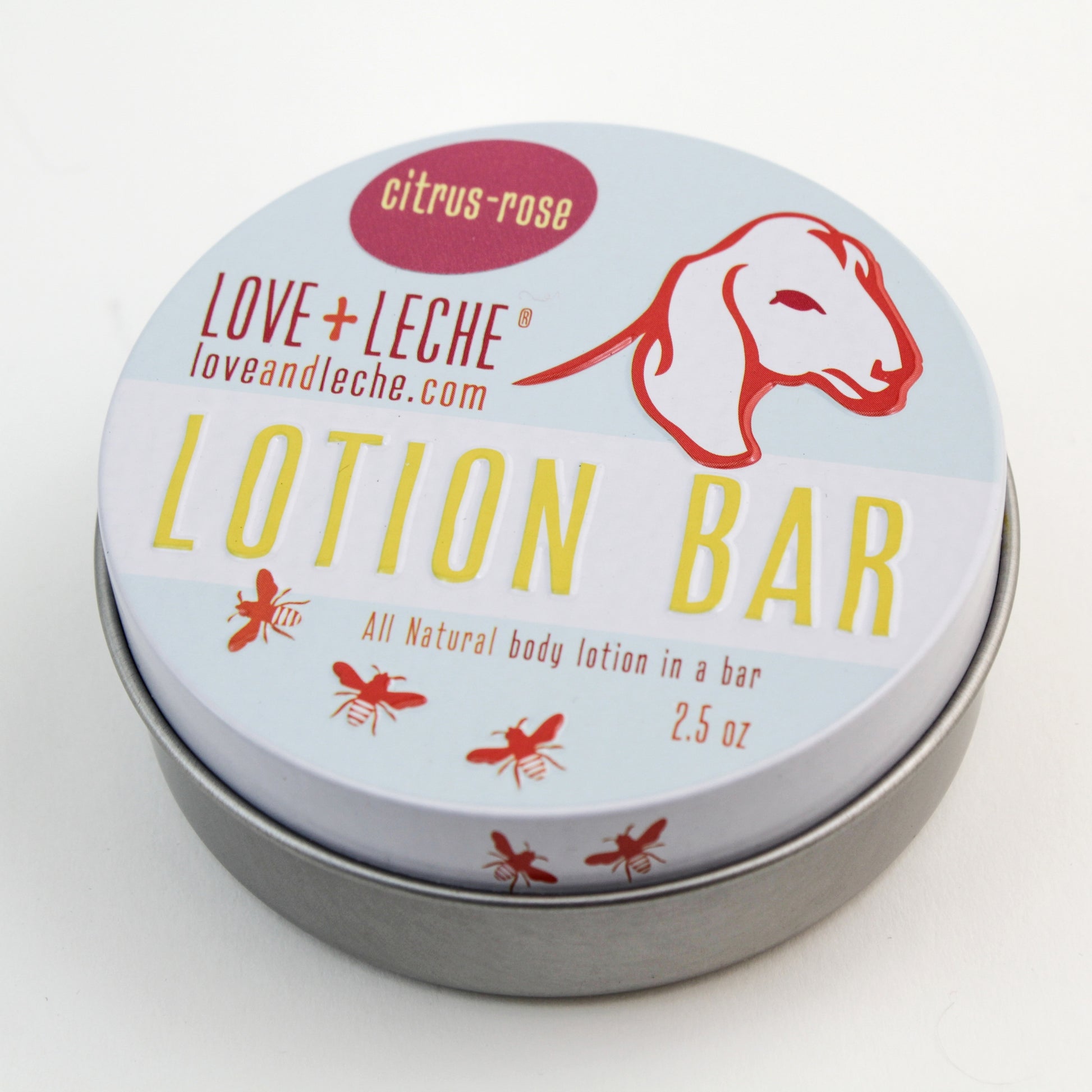 A circular tin container of Love + Leche Lotion Bar in the citrus-rose scent. The white lid is adorned with a red goat illustration and product details, including the website "loveandleche.com." The label also showcases small red bees and emphasizes the benefits of beeswax for deeply moisturized skin.