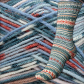 A single knit sock featuring intricate Fair Isle patterns in shades of green, red, and white rests against a textured backdrop of tangled yarn in blue, green, and red hues. The colorful and detailed background enhances the rich design of the Regia 4-ply Design Line by Arne & Carlos from Sirdar - Regia, Schachenmayr & Rowan.