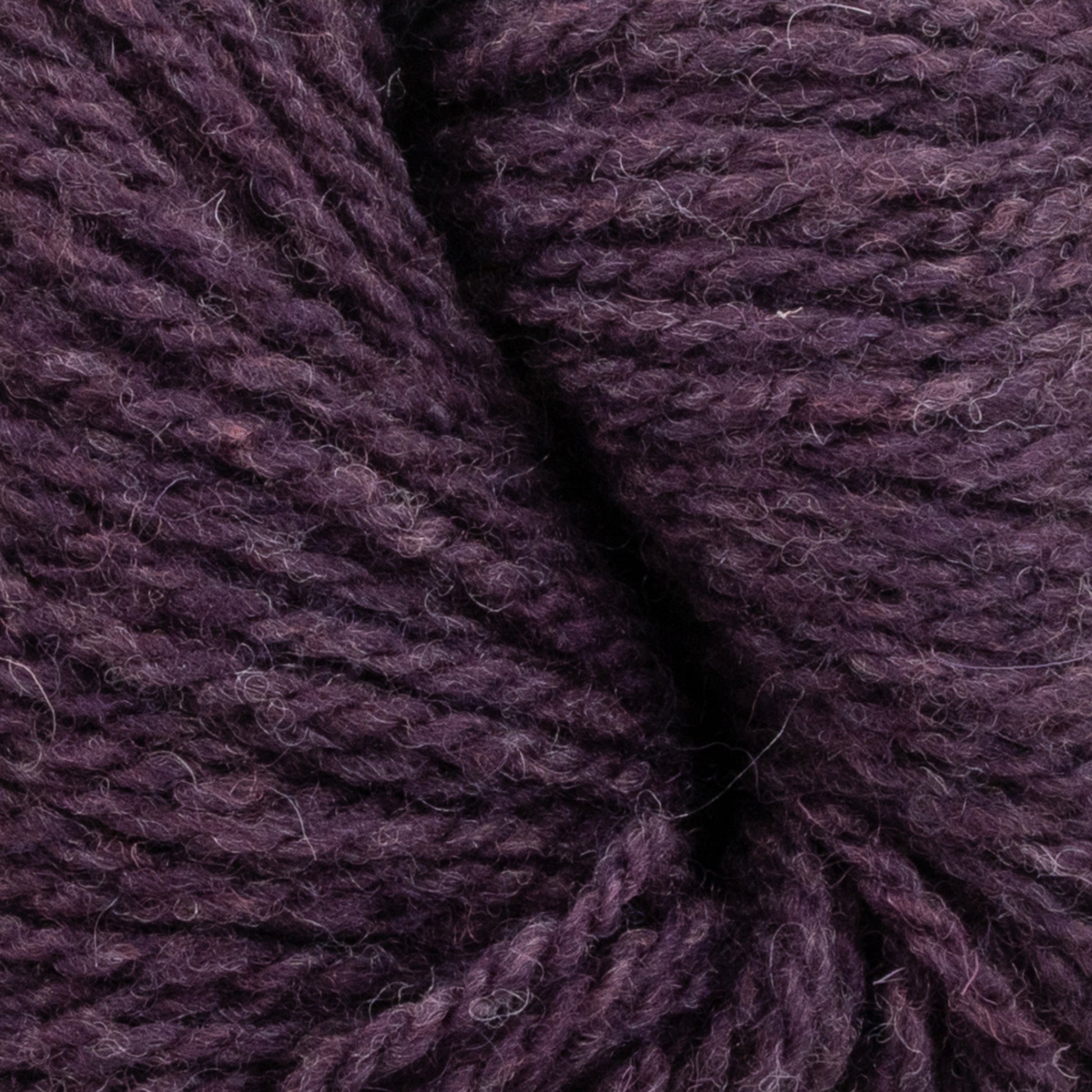 Close-up image of dark purple Patagonia Organic Merino by Juniper Moon Farm yarn from Knitting Fever / Euro Yarns, showcasing its texture and fibers in detail. The sustainable yarn features a coarse, slightly fuzzy appearance with multiple twisted strands. The rich color exhibits slight tonal variations throughout the coils.