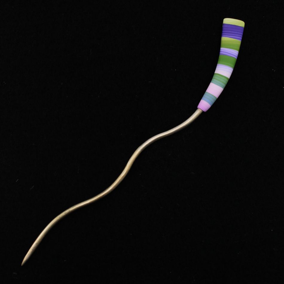 The Striped Color Stick Shawl Pin by Bonnie Bishoff Designs, created by the renowned Bonnie Bishoff, features a brass bodkin with a long, slender, wavy body. Its handle is wrapped in vibrant thread bands of purple, blue, green, and yellow against a black background. This intricate metal design combines both functionality and decorative elegance.