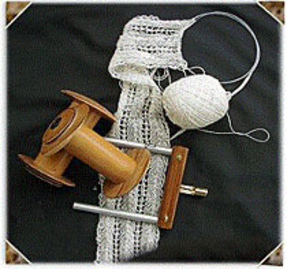 A handmade white knitted scarf is in progress on circular knitting needles, with a ball of white yarn attached. Beside it, an empty hand-turned bobbin from the Majacraft Lace Kit for Majacraft Wheels by Majacraft Co. is placed on a black surface.