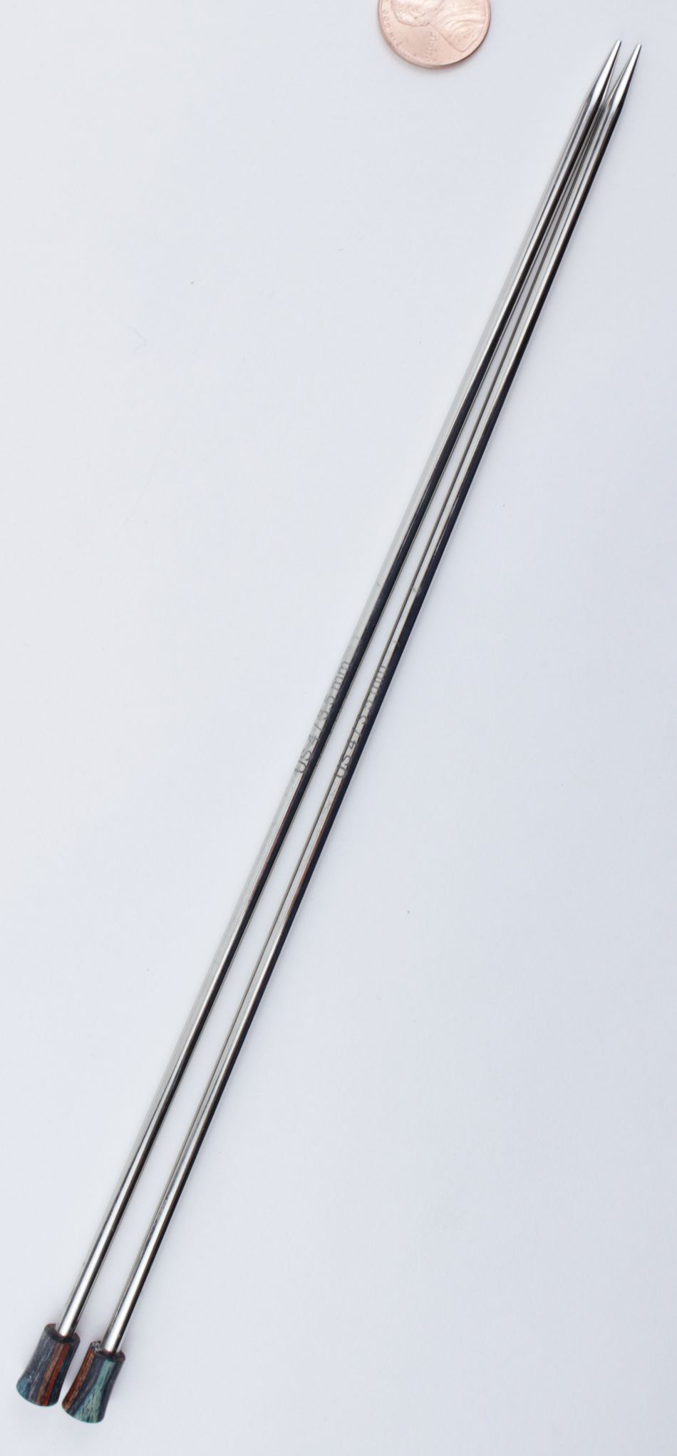 Two long, thin Accessories Unlimited Nova Platina Single Point Knitting Needles with tapered, pointed tips are placed parallel to each other on a light surface. Near the top edge of the image, a small copper coin provides a sense of scale.