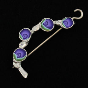 The Spring Bud Shawl Pin by Bonnie Bishoff Designs showcases an elegant silver brooch adorned with intricate details, featuring four round enamel sections, each depicting a purple rosebud on a green stem. Ideal for knit wearables, the brooch includes a secure locking pin on the back. The black background beautifully highlights its intricate elegance.