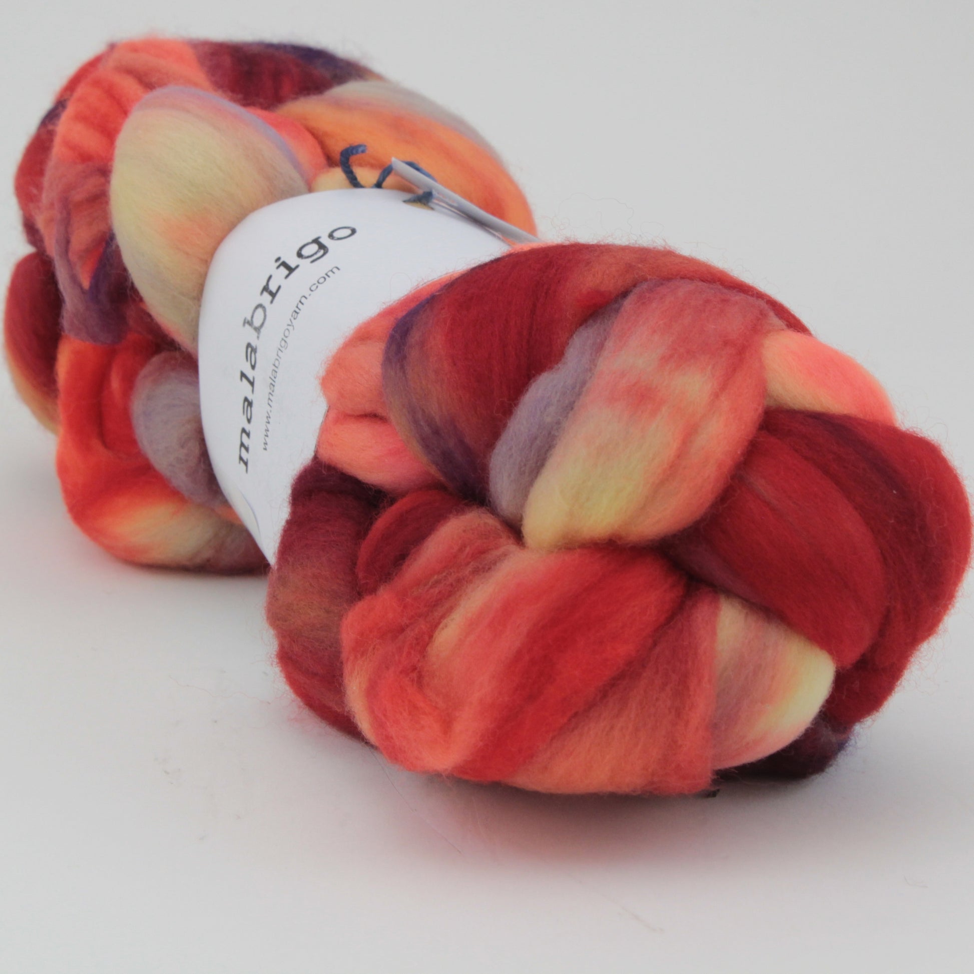 A skein of Nube by Malabrigo features hand-dyed, luxurious merino wool roving in vibrant hues including red, orange, yellow, and purple. The wool is braided for a thick, textured appearance and is wrapped with a white label around the middle.