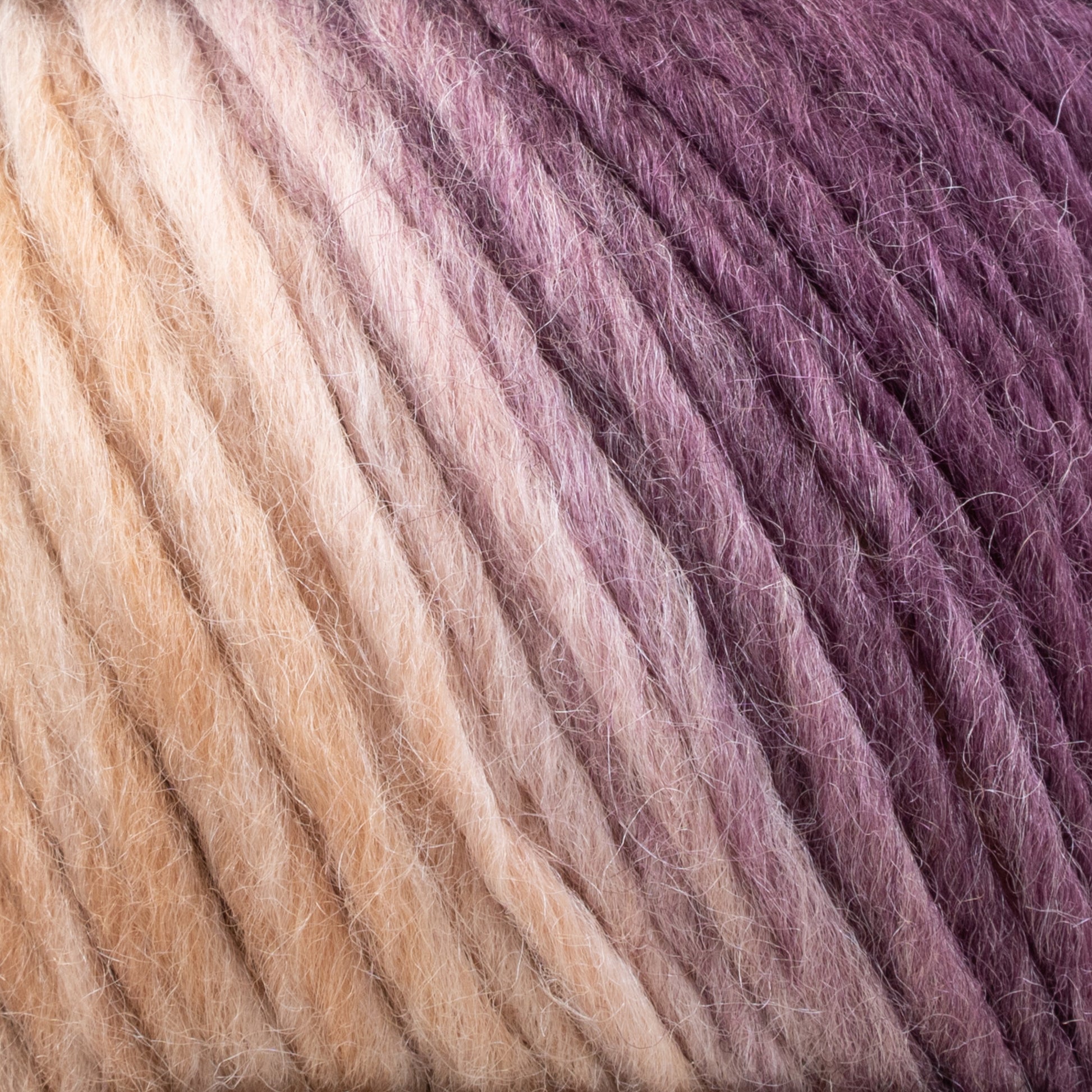 Close-up of Louisa Harding Amitola Grande by Knitting Fever / Euro Yarns, featuring a self-striping, multi-colored yarn with a smooth gradient from beige to purple. The texture appears soft and slightly fuzzy, suggesting it's made of wool or a similar material. The colors blend seamlessly to create a rich ombré effect.