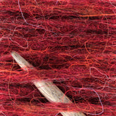 Close-up of a wooden crochet hook nestled in a tangle of Plötulopi Unspun Icelandic Wool Yarn. The Berroco, Inc. wool displays varying shades of red intertwined with strands of orange, pink, and purple, creating a richly textured appearance. The hook is partially visible as it emerges from the vibrant yarn.