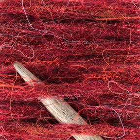 Close-up of a wooden crochet hook nestled in a tangle of Plötulopi Unspun Icelandic Wool Yarn. The Berroco, Inc. wool displays varying shades of red intertwined with strands of orange, pink, and purple, creating a richly textured appearance. The hook is partially visible as it emerges from the vibrant yarn.