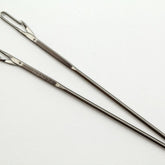 Two long, silver darning needles with latch hook eyes and pointed ends are arranged parallel on a white surface. The needle on the left, designed for easy threading, has the brand name "Accessories Unlimited" engraved near the eye, while the needle on the right has no visible engravings.