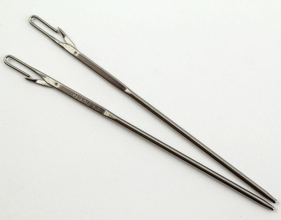 Two long, silver darning needles with latch hook eyes and pointed ends are arranged parallel on a white surface. The needle on the left, designed for easy threading, has the brand name "Accessories Unlimited" engraved near the eye, while the needle on the right has no visible engravings.