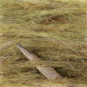 Close-up of a weaving project with thick, light yellow-green fibers from the Plötulopi Unspun Icelandic Wool Yarn by Berroco, Inc. The fibers are tightly intertwined, with a wooden weaving needle partially visible at an angle emerging from them. The texture of the Icelandic wool and the needle is clearly detailed.