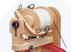 The Schacht Bulky Plyer Flyer Package, produced by Schacht Spindle Co., features a large spool of white fiber mounted on a wooden spinning wheel. The components are crafted from light-colored wood and complemented by black metal hooks, exhibiting a modern design aesthetic. The image prominently captures the upper section, showcasing the spool and its support structure.