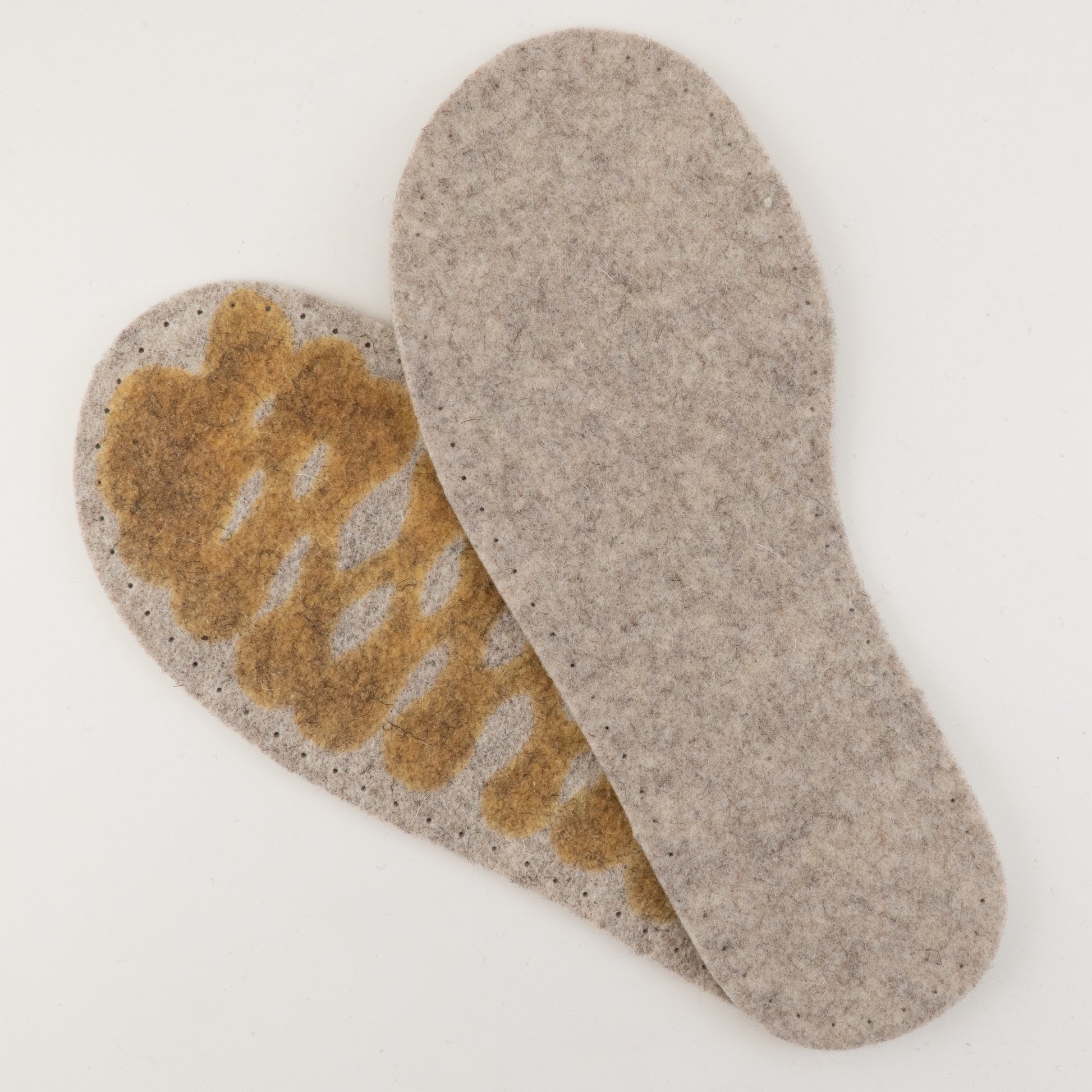 A pair of Joe's Toes Adult's Thick Felt Slipper Soles w/Latex Grip in grey is displayed on a white background. The left insole, made from eco-friendly wool, shows a worn, brownish area indicating high wear and use, while the right insole remains relatively clean and unworn.