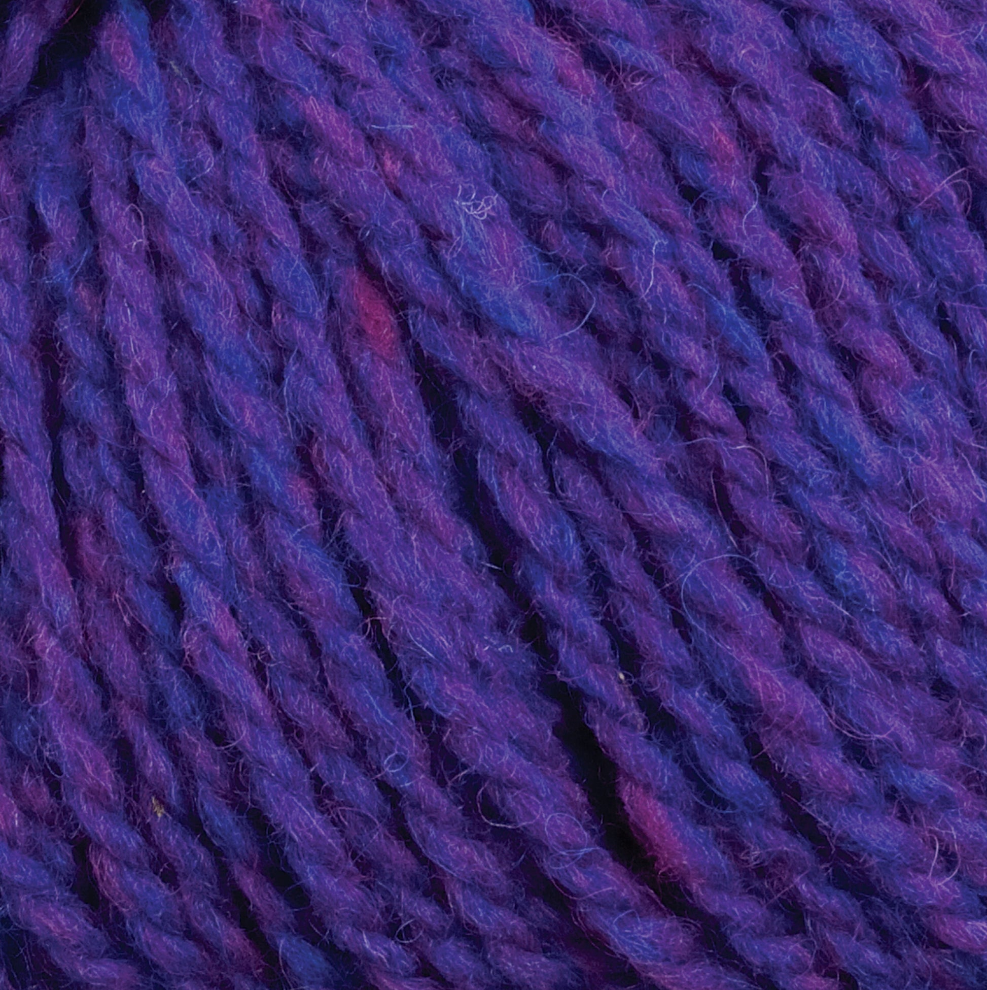 A close-up image of the Harrisville Designs Shetland Yarn - Unwashed Cones reveals thick, purple intertwined strands of fiber, blending shades of purple and blue with hints of red. The texture appears soft and suitable for lightweight blankets or Fair Isle knitting designs.