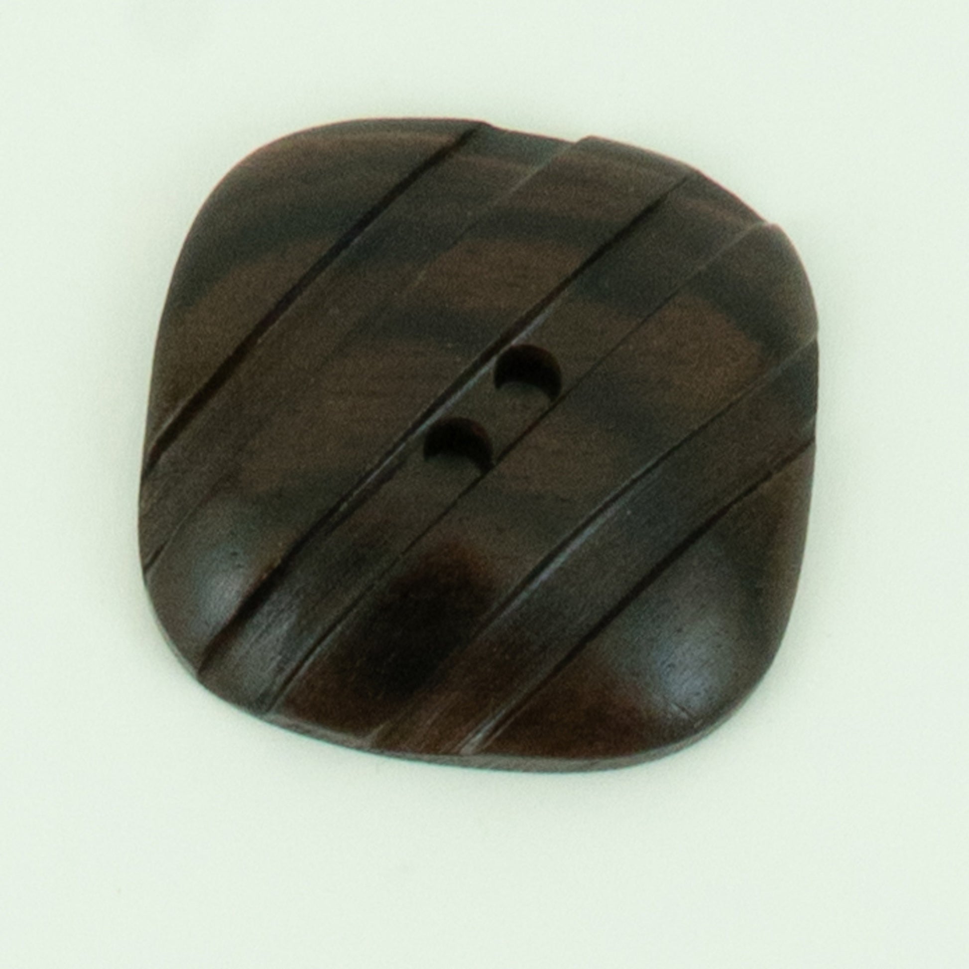 A close-up image of the Wood Grooved Square Button by Buttons Etc. showcases its square, dark brown wooden design with slightly rounded edges. The button features two holes and diagonal grooves that create a striped pattern, measuring 1 3/8" in diameter against a plain white background.