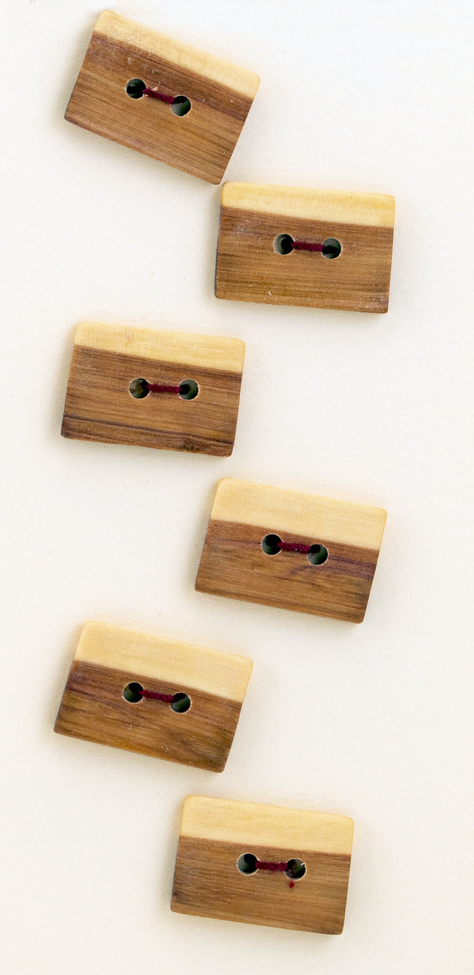 An arrangement of Six Square or Oblong Buttons by Favour Valley Woodworking, each handcrafted from local wood. These rectangular buttons with rounded edges feature two pairs of circular holes connected by short red threads and are aligned diagonally in two columns against a white background for a unique touch.