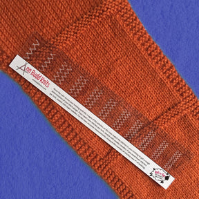 A close-up of an orange knitted fabric laid on a blue surface. A transparent stitch gauge ruler, labeled "The Ann Budd Stitch Gauge Ruler" from Ann Budd, is placed horizontally across the material, offering accurate measurement in both inches and centimeters. This essential knitting accessory ensures precise dimensions for your projects.