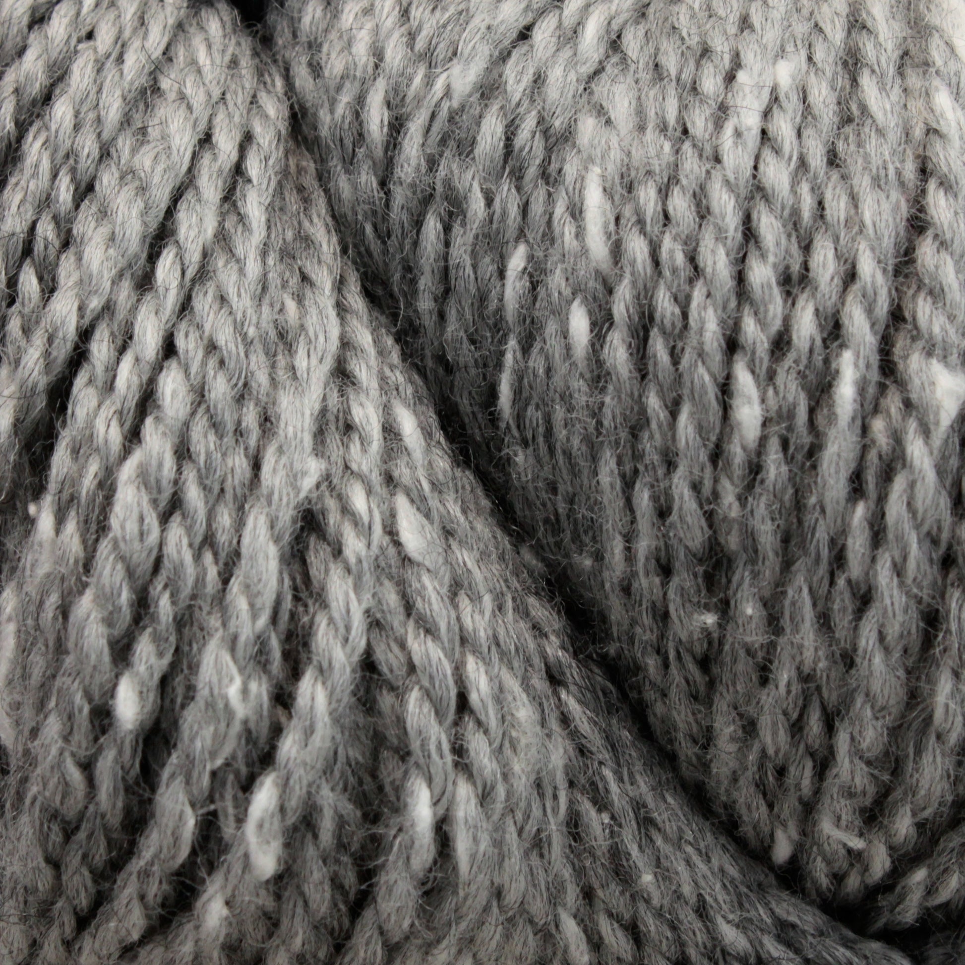 Close-up image of twisted, thick, grey Acadia by The Fibre Co. (UK) Ltd yarn with flecks of white. The texture appears soft and fuzzy, highlighting the individual strands and twists of the yarn, creating a subtle tweed effect suggestive of a cozy material ideal for knitting or crocheting projects.