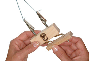 Two hands are holding the Leclerc Double Fringe Twister from Leclerc Looms, a wooden jig featuring two alligator clips attached to wires. One hand is holding the base of the twister, while the other is adjusting a part. The jig has a logo with a spiral design burned into the wood.