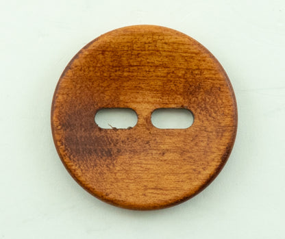 The "Wood Foxeye" by Buttons Etc. is a round wooden button with a 1 3/8" diameter and a smooth texture, featuring a reddish-brown finish. This two-hole button has elongated, horizontal holes in the center for sewing. The surface shows slight variations in color and grain, highlighting its natural wood material.