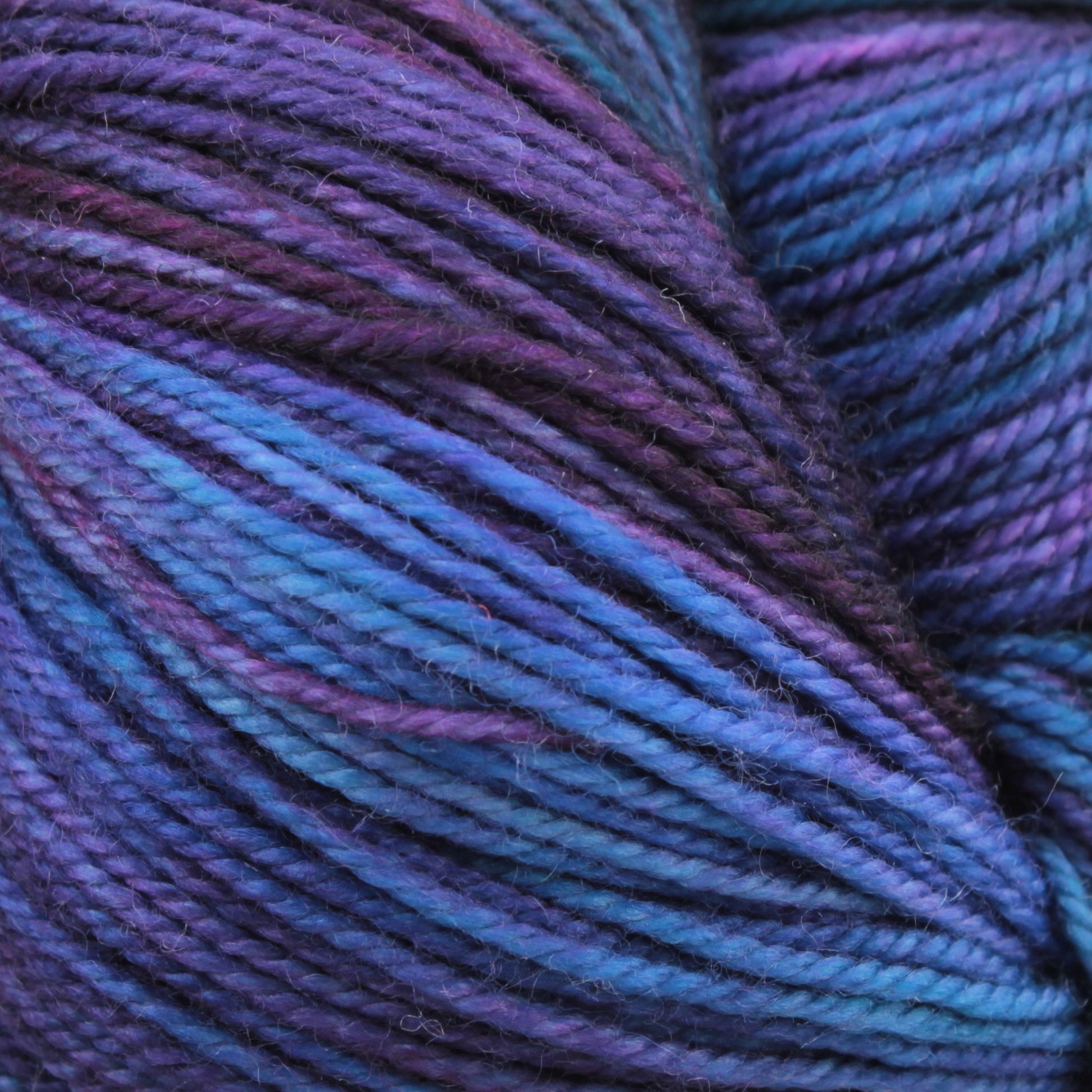 Close-up of purple, blue, and teal colored strands of Malabrigo Sock twisted together. The texture of this kettle-dyed multicolors yarn from Malabrigo Yarn is soft and slightly fuzzy, with varying shades blending smoothly from one color to another, creating a rich, vibrant appearance.