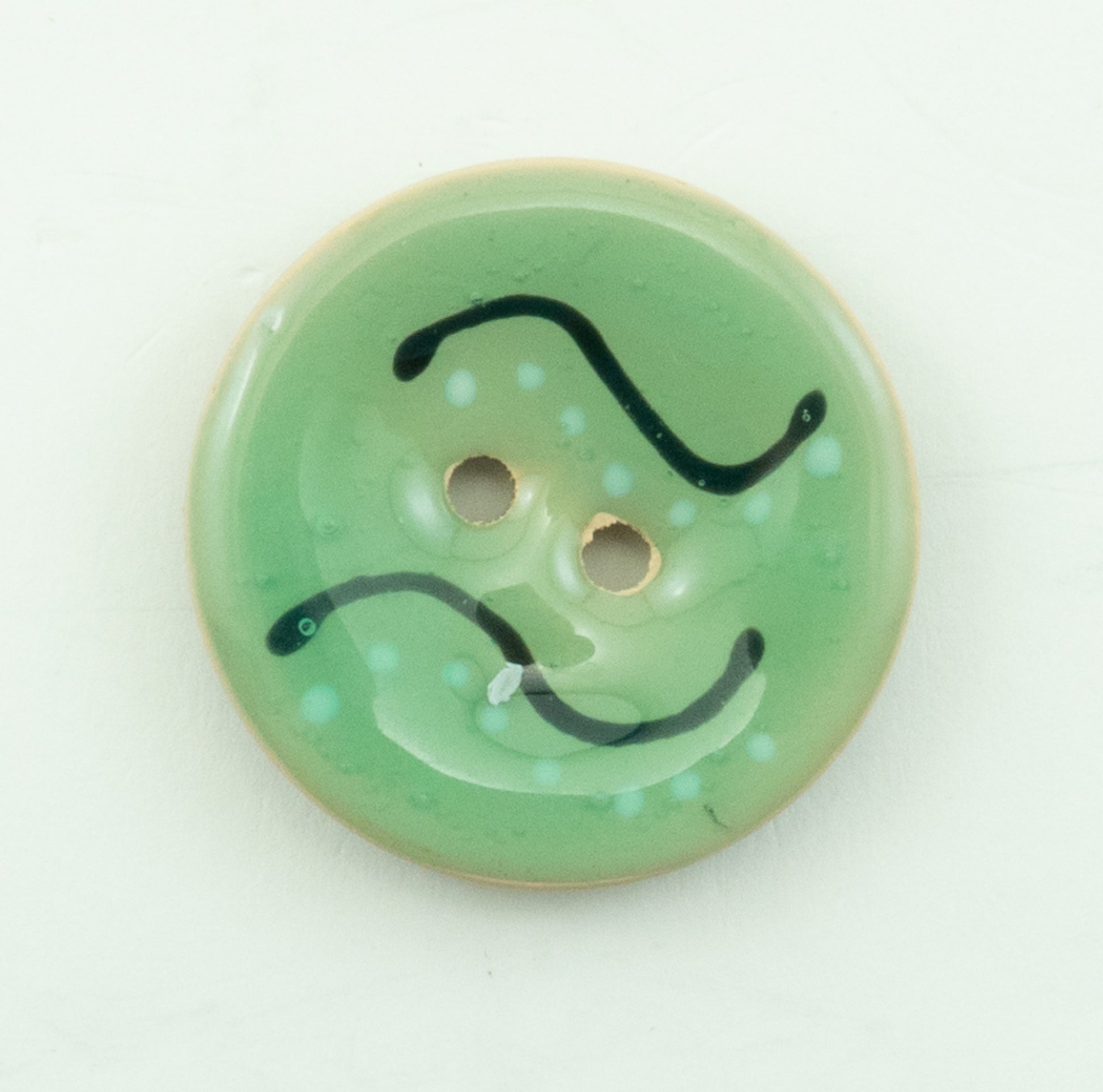 A close-up of the Coco Enamel Fa la la Button by Buttons Etc. This round, green button with a 1 1/8" diameter showcases two holes in the center. It features a light beige rim and an abstract black and light blue squiggly line pattern on its green surface.