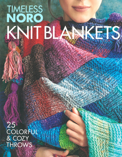 A person wrapped in a vibrant, multicolored knit blanket. The text on the image reads: "Timeless Noro: Knit Blankets: 25 Colorful & Cozy Throws" from Knitting Fever / Euro Yarns. The person's face is partially visible, and they are holding the edge of the cozy throw.