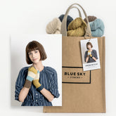 A brown paper bag labeled "Blue Sky Fibers" filled with colorful yarns. Attached is a tag featuring a photo of a woman in a striped dress, wearing warm baby alpaca mittens in blue, white, yellow, and brown. The tag reads "Blue Sky Fibers Virginia Mitts Kit.