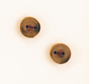 Two handmade Round or Square Buttons - Mixed Woods by Favour Valley Woodworking, showcasing visible tree rings, are positioned diagonally on a plain, off-white background. Each button has two holes with red thread stitched through them. The natural texture and grain of the local wood are clearly visible.