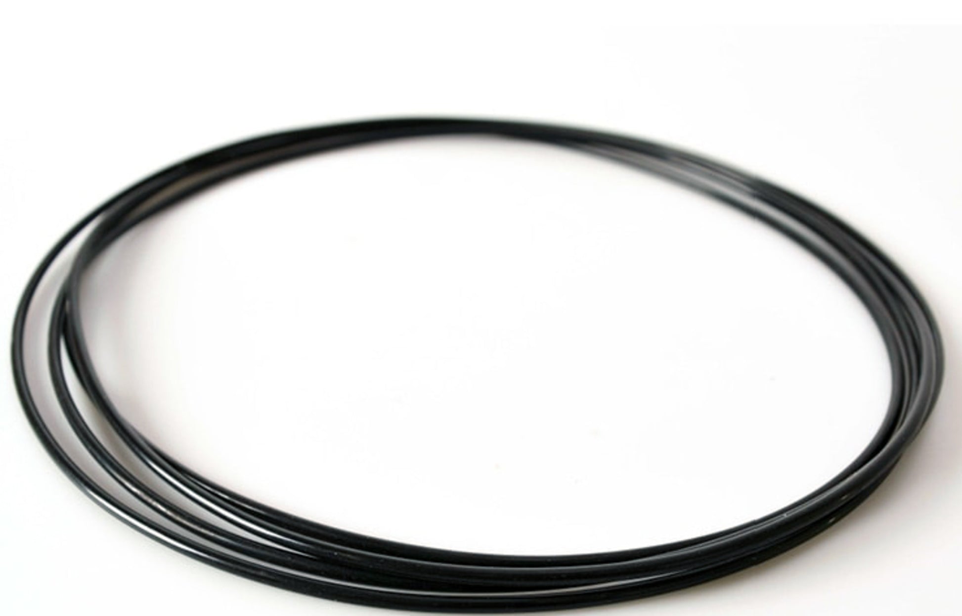 A Schacht Black Poly Drive Band from Schacht Spindle Co. lies coiled on a white surface, forming a nearly perfect circle with slight overlaps at the ends. The band appears flexible and smooth, reminiscent of those used in electronics or other technical applications.