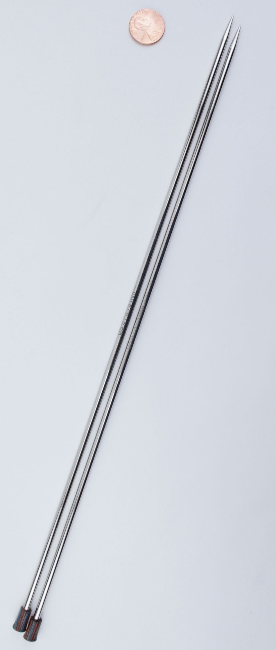 Two long, smooth, and shiny skewers with pointed tips are placed side by side on a light surface. There is a penny at the top for scale. Resembling the sleek design of Accessories Unlimited's Nova Platina Single Point Knitting Needles, they feature small black handles on one end and appear almost like hollow brass pipes.