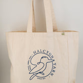 The Halcyon Yarn Recycled Cotton Tote Bag w/Pocket, a beige tote crafted from recycled cotton canvas, proudly displays a simplistic blue bird logo along with the text "Halcyon Yarn" in a circular design that reads "Since 1971" and "Fine Fiber Tools." Hanging gracefully from one's hand, an "ECO" tag is visible on the side, making this oversized canvas tote both stylish and sustainable.